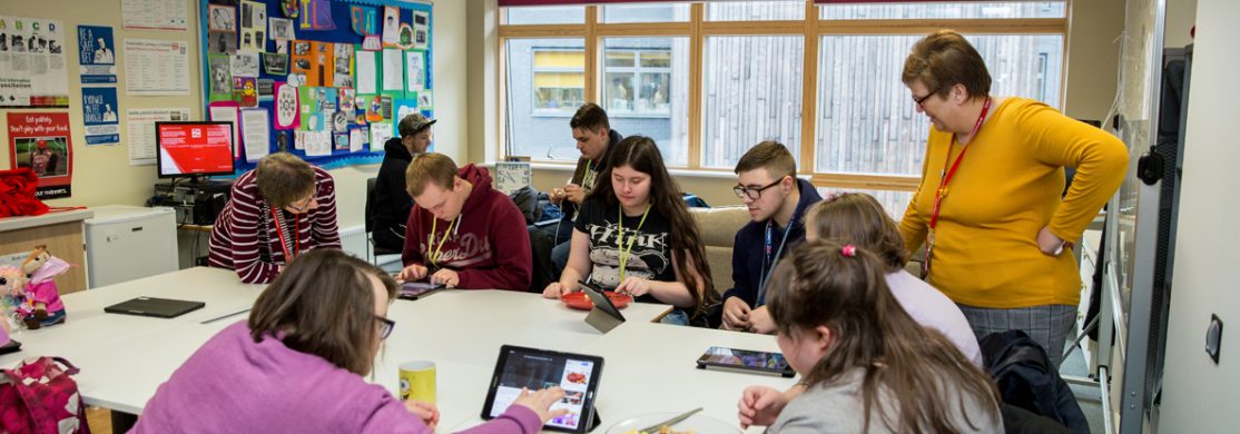 Group of students sat around a table enjoying a snack and using tablets