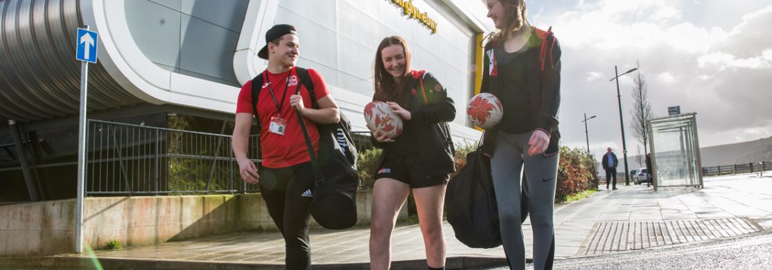 3 students holding rugby balls and sports kit in front of sports building
