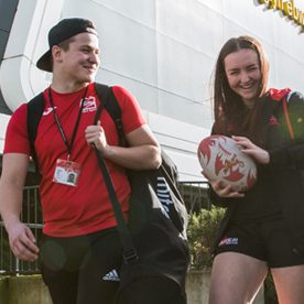 male and female student carry rugby ball and sports bags